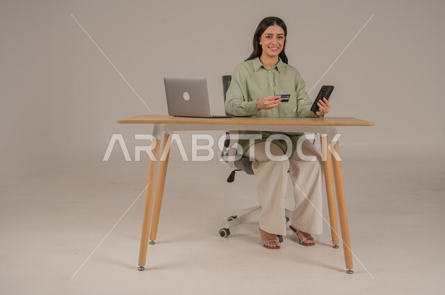الشراء والدفع عن طريق بطاقة الصراف الإئتمانية، سهولة تسديد الفواتير عبر الحساب البنكي، بورتريه لامراة عربية خليجية سعودية ترتدي الزي الكاجوال تجلس على طاولة المكتب تحمل بيدها كرت البنك المصرفي والهاتف المحمول، صورة لكامل الجسم، خلفية رمادية