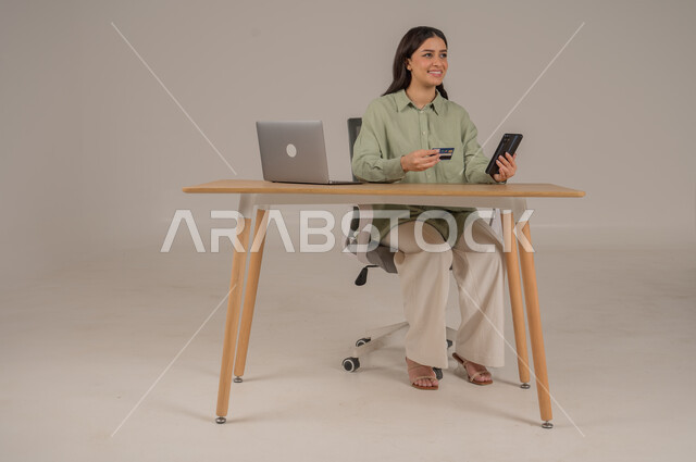 سهولة تسديد الفواتير عبر الحساب البنكي، الشراء والدفع عن طريق بطاقة الصراف الإئتمانية، بورتريه لامراة عربية خليجية سعودية ترتدي الزي الكاجوال تجلس على طاولة المكتب تحمل بيدها كرت البنك المصرفي والهاتف المحمول، صورة لكامل الجسم، خلفية رمادية