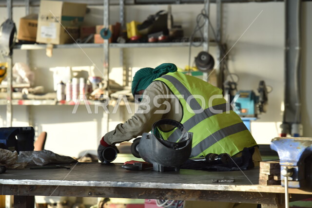 تجهيز وتوصيل الحديد بمعدات وأدوات لحامية خاصة، متابعة سير العمل في ورش الحدادة بمهارة وإتقان، ارتداء الزي الواقي للحماية من الشرار المتناثر في المعمل, عامل عربي خليجي سعودي يرتدي قناع الحماية والقفازات يقوم بلحام المعدن وصهره, وظائف ومهن صناعية للشباب السعودي