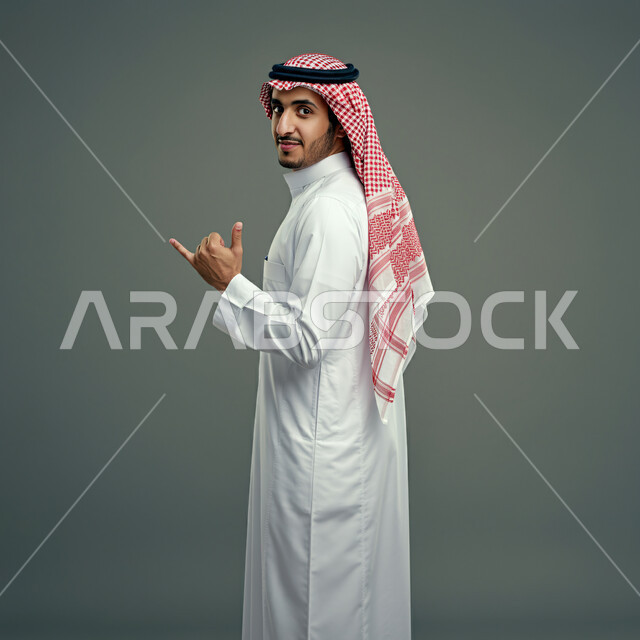 الثقة بالنفس والشعور بالقوة، بورتريه من الخلف لشاب عربي خليجي سعودي يرتدي الثوب التقليدي والشماغ ينظر إلى الكاميرا، إيماءات يدين مختلفة، مفهوم الرجولة والأناقة، الاهتمام بالمظهر الخارجي، خلفية رمادية