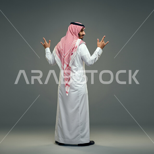 الاهتمام بالمظهر الخارجي، الثقة بالنفس والشعور بالقوة، بورتريه من الخلف لشاب عربي خليجي سعودي يرتدي الثوب التقليدي والشماغ يقف باستقامة، إيماءات يدين مختلفة، مفهوم الرجولة والأناقة، صورة الجسم بالطول الكامل، خلفية رمادية