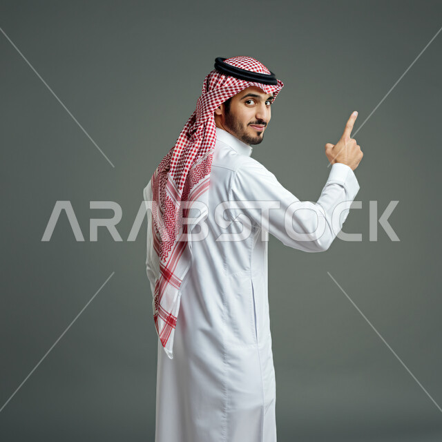 الثقة بالنفس والشعور بالقوة، بورتريه من الخلف لشاب عربي خليجي سعودي يرتدي الثوب التقليدي والشماغ يرفع أصبع السبابة يشير إلى شيء ما ينظر إلى الكاميرا، مفهوم الرجولة والأناقة، الاهتمام بالمظهر الخارجي، خلفية رمادية
