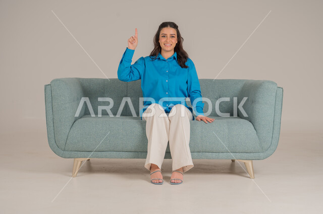 النظر إلى الكاميرا بإيماءات السعادة والسرور، بورتريه لشابة عربية خليجية سعودية ترتدي الزي الكاجوال تجلس على كنبة مريحة تشير باصبع السبابة نحو الأعلى، الإشارة بالاصابع نحو اتجاه محدد، الأناقة والاهتمام بالموضة والمظهر الخارجي، الجسم بالطول الكامل، خلفية رمادية