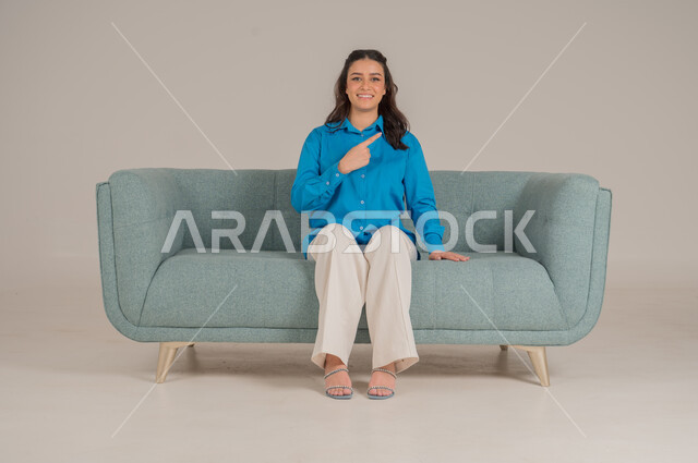 النظر إلى الكاميرا بإيماءات السعادة والسرور، بورتريه لشابة عربية خليجية سعودية ترتدي الزي الكاجوال تجلس على كنبة مريحة تشير باصبع السبابة نحو الأعلى، الإشارة بالاصابع نحو اتجاه محدد، الأناقة والاهتمام بالموضة والمظهر الخارجي، الجسم بالطول الكامل، خلفية رمادية