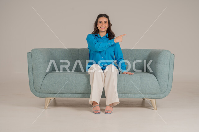 Looking at the camera with gestures of happiness and pleasure, portrait of a young Saudi Arabian Gulf woman wearing casual clothes sitting on a comfortable sofa pointing her index finger upwards, pointing fingers in a specific direction, elegance, interest in fashion and appearance, full body, gray background