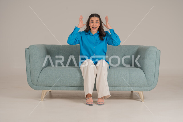 المرح وقضاء أوقات ممتعة، رفع اليدين بحركات يدين تشير إلى السخرية والضحك، بورتريه لشابة عربية خليجية سعودية ترتدي الزي الكاجوال تجلس على كنبة مريحة في غرفة المعيشة وتضحك، الأناقة والاهتمام بالموضة والمظهر الخارجي، الجسم بالطول الكامل، خلفية رمادية