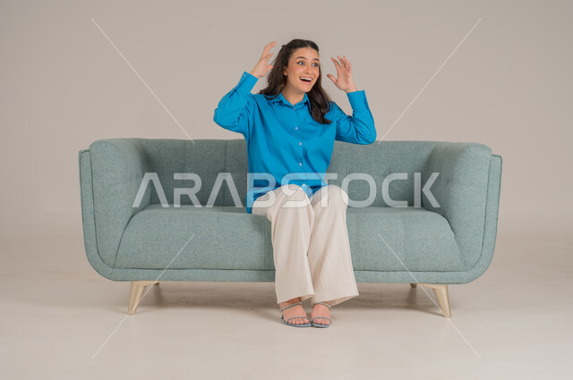 المرح وقضاء أوقات ممتعة، رفع اليدين بحركات يدين تشير إلى السخرية والضحك، بورتريه لشابة عربية خليجية سعودية ترتدي الزي الكاجوال تجلس على كنبة مريحة في غرفة المعيشة وتضحك، الأناقة والاهتمام بالموضة والمظهر الخارجي، الجسم بالطول الكامل، خلفية رمادية