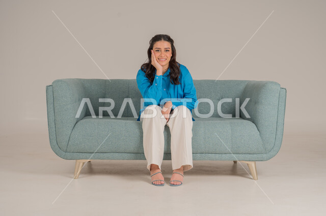 وضع اليد على الخد بإيماءات الملل، شرود الذهن والتفكير العميق، بورتريه لشابة عربية خليجية سعودية ترتدي الزي الكاجوال تجلس على كنبة مريحة في غرفة المعيشة وتشعر بالضجر، مواكبة صيحات الموضة، الأناقة والاهتمام المظهر الخارجي، الجسم بالطول الكامل، خلفية رمادية