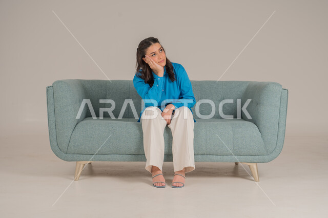 وضع اليد على الخد بإيماءات الملل، شرود الذهن والتفكير العميق، بورتريه لشابة عربية خليجية سعودية ترتدي الزي الكاجوال تجلس على كنبة مريحة في غرفة المعيشة وتشعر بالضجر، مواكبة صيحات الموضة، الأناقة والاهتمام المظهر الخارجي، الجسم بالطول الكامل، خلفية رمادية