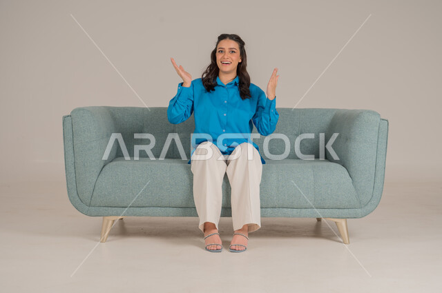 الإشارة بالاصابع نحو اتجاه محدد، النظر إلى الكاميرا بإيماءات السعادة والسرور، بورتريه لشابة عربية خليجية سعودية ترتدي الزي الكاجوال تجلس على كنبة مريحة تشير باصبيعها السبابة نحو الأعلى، الأناقة والاهتمام بالموضة والمظهر الخارجي، الجسم بالطول الكامل، خلفية رمادية