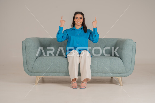 Pointing fingers in a specific direction, looking at the camera with gestures of happiness and pleasure, portrait of a young Saudi Arabian Gulf woman in casual wear sitting on a comfortable sofa pointing her index finger upwards, elegance and interest in fashion and appearance, full body, gray background
