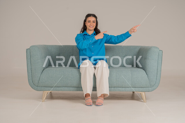 الإشارة بالاصابع نحو اتجاه محدد، النظر إلى الكاميرا بإيماءات السعادة والسرور، بورتريه لشابة عربية خليجية سعودية ترتدي الزي الكاجوال تجلس على كنبة مريحة تشير باصبيعها السبابة نحو الأعلى، الأناقة والاهتمام بالموضة والمظهر الخارجي، الجسم بالطول الكامل، خلفية رمادية