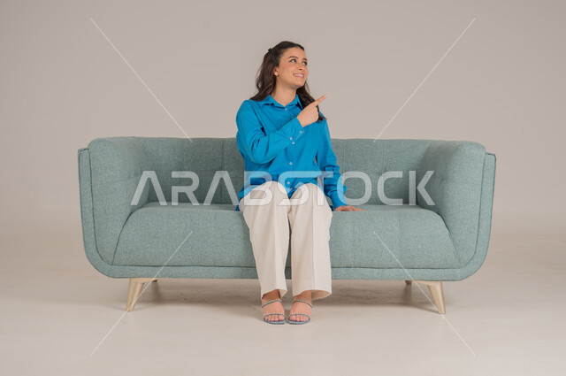 Pointing towards a specific direction with gestures of pleasure and cheerfulness, portrait of a young Saudi Arabian Gulf woman wearing casual clothes sitting on a comfortable sofa pointing her index finger upwards, elegance and interest in fashion and appearance, full body, gray background