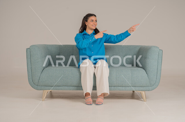 الإشارة نحو اتجاه محدد بإيماءات السرور والبشاشة، بورتريه لشابة عربية خليجية سعودية ترتدي الزي الكاجوال تجلس على كنبة مريحة تشير بيديها نحو الأعلى، الأناقة والاهتمام بالموضة والمظهر الخارجي، الجسم بالطول الكامل، خلفية رمادية