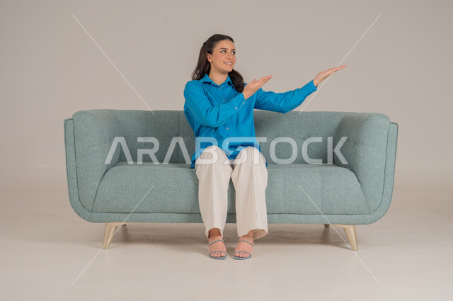 الإشارة نحو اتجاه محدد بإيماءات السرور والبشاشة، بورتريه لشابة عربية خليجية سعودية ترتدي الزي الكاجوال تجلس على كنبة مريحة تشير بيديها نحو الأعلى، الأناقة والاهتمام بالموضة والمظهر الخارجي، الجسم بالطول الكامل، خلفية رمادية