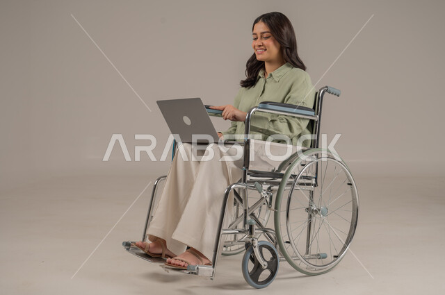 Using modern and advanced technologies, accomplishing work tasks remotely, library professions and jobs, portrait of a seated Saudi Arabian Gulf woman wearing casual clothes sitting on a wheelchair working on a laptop, full body image, gray background