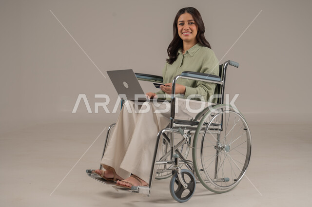 سهولة تسديد الفواتير عبر الحساب البنكي وتطبيقات الحاسوب المحمول، الشراء والدفع عن طريق بطاقة الصراف الإئتمانية، بورتريه لامراة عربية خليجية سعودية مقعد ترتدي الزي الكاجوال تجلس على كرسي متحرك تحمل بيدها كرت البنك المصرفي، صورة لكامل الجسم، خلفية رمادية