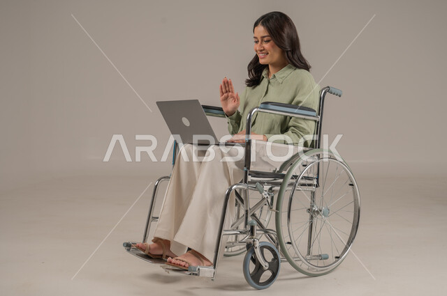 اجراء مكالمات فيديو، التواصل مع الاهل والاصدقاء، انجازمهام العمل عن بعد، عم المجتمع السعودي لذوي الهمم، بورتريه لامراة عربية خليجية سعودية مقعد ترتدي الزي الكاجوال تجلس على كرسي متحرك تعمل على جهاز الحاسوب المحمول، صورة لكامل الجسم، خلفية رمادية