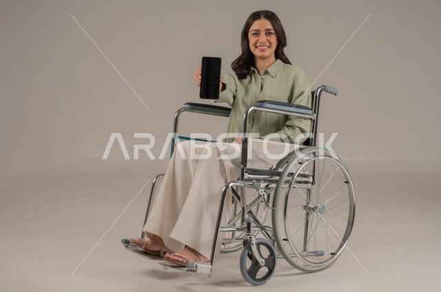 مهن ووظائف مكتبة، عرض شاشة سوداء فارغة، انجاز العمل عن بعد، دعم المجتمع السعودي لذوي الهمم، بورتريه لامراة عربية خليجية سعودية مقعد ترتدي الزي الكاجوال تجلس على كرسي متحرك تحمل بيدها الهاتف المحمول، صورة لكامل الجسم، خلفية رمادية