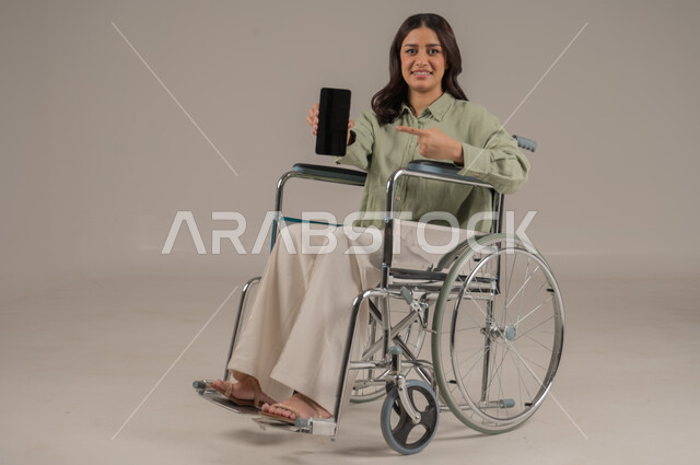 عرض شاشة سوداء فارغة، انجاز العمل عن بعد، مهن ووظائف مكتبة، دعم المجتمع السعودي لذوي الهمم، بورتريه لامراة عربية خليجية سعودية مقعد ترتدي الزي الكاجوال تجلس على كرسي متحرك تشير باصبع السبابة الى الهاتف المحمول، صورة لكامل الجسم، خلفية رمادية