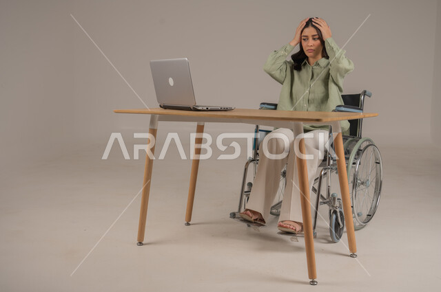 الشعور بالانزعاج والتوتر من سماع الأخبار السيئة والمزعجة، تعابير الدهشة والذهول، بورتريه لامراة عربية خليجية سعودية مقعدة ترتدي الزي الكاجوال تجلس على طاولة المكتب تضع يديها على رأسها بإيماءات الصدمة والمفاجأة، اسلوب حياة ذوي الهمم، صورة لكامل الجسم، خلفية رمادية