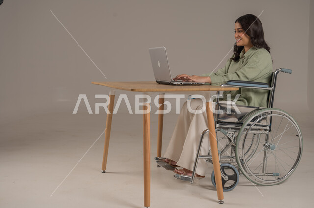 Completing work tasks remotely, using modern and advanced technologies, office jobs and professions, Saudi society for people with disabilities, side portrait of a disabled Saudi Arabian Gulf woman wearing casual clothes sitting at an office table working on a computer, full body image, gray background