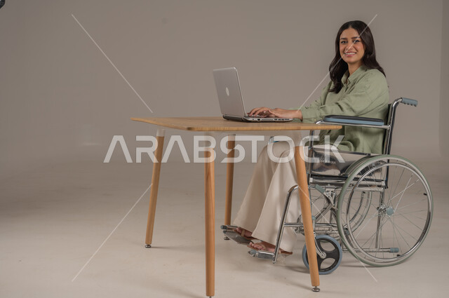 استخدام التقنيات الحديثة والمتطورة، انجاز مهام العمل عن بعد، مهن ووظائف مكتبية، عم المجتمع السعودي لذوي الهمم، بورتريه من الجانب لامراة عربية خليجية سعودية مقعدة ترتدي الزي الكاجوال تجلس على طاولة المكتب تعمل على جهاز الحاسوب، صورة لكامل الجسم، خلفية رمادية
