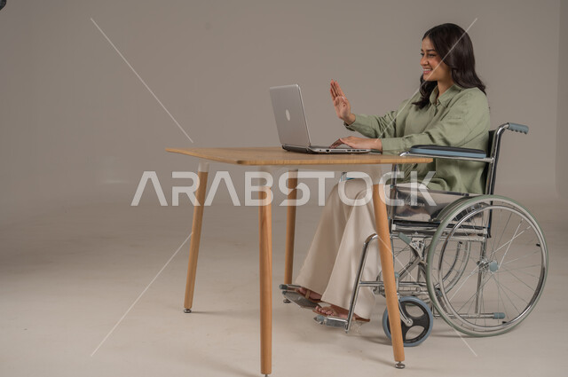 اجراء مكالمات فيديو، التواصل مع الاهل والاصدقاء، انجازمهام العمل عن بعد، عم المجتمع السعودي لذوي الهمم، بورتريه من الجانب لامراة عربية خليجية سعودية مقعدة ترتدي الزي الكاجوال تجلس على طاولة المكتب تعمل على جهاز الحاسوب، صورة لكامل الجسم، خلفية رمادية