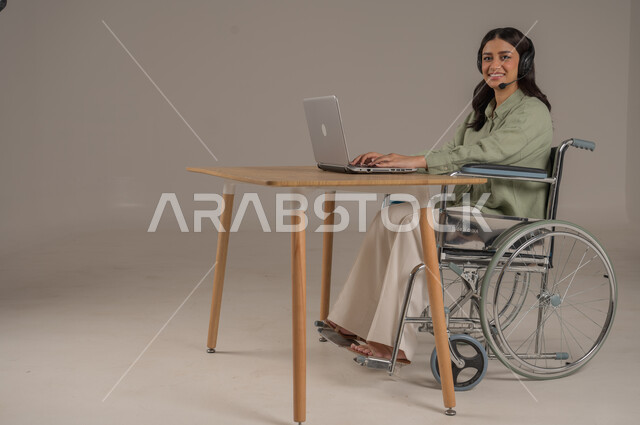 الرد والاجابة على الاستفسارات، العمل في مجال خدمة العملاء، دعم المجتمع السعودي لذوي الهمم، بورتريه من الجانب لامراة عربية خليجية سعودية مقعدة ترتدي الزي الكاجوال تجلس على طاولة المكتب تعمل على جهاز الحاسوب، صورة لكامل الجسم، خلفية رمادية