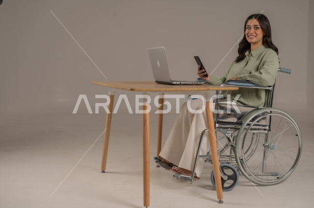 Saudi community support for people with disabilities, remote work, library jobs and professions, use of modern and advanced technologies, side portrait of a disabled Saudi Arabian Gulf woman wearing casual clothes sitting at an office table holding a mobile phone in her hand, full body image, gray background