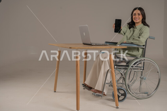 عرض شاشة سوداء فارغة، دعم المجتمع السعودي لذوي الهمم، مهن ووظائف مكتبية، استخدام التقنيات الحديثة والمتطورة، بورتريه من الجانب لامراة عربية خليجية سعودية مقعدة ترتدي الزي الكاجوال تجلس على طاولة المكتب تحمل بيدها الهاتف المحمول، صورة لكامل الجسم، خلفية رمادية