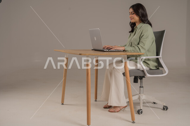 استخدام التقنيات الحديثة والمتطورة، انجاز مهام العمل عن بعد، مهن ووظائف مكتبية، بورتريه من الجانب لامراة عربية خليجية سعودية ترتدي الزي الكاجوال تجلس على طاولة المكتب تعمل على جهاز الحاسوب المحمول، صورة لكامل الجسم، خلفية رمادية