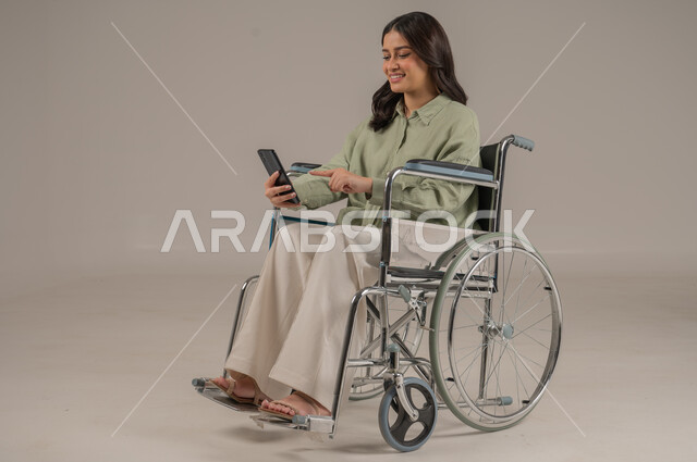 استخدام الاجهزة التقنية الحديثة، تعابير السرور والسعادة، الدردشة والتواصل مع الاهل والاصدقاء، بورتريه لامراة عربية خليجية سعودية مقعد ترتدي الزي الكاجوال تجلس على كرسي متحرك تشير باصبع السبابة الى الهاتف المحمول، صورة لكامل الجسم، خلفية رمادية