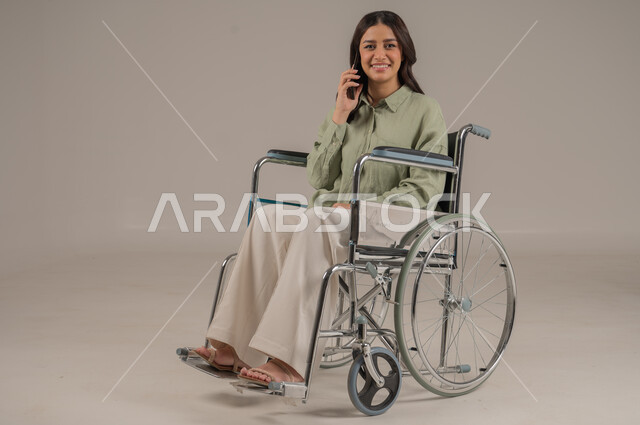 اجراء المكالمات الهاتفية، استخدام الاجهزة التقنية الحديثة، الدردشة والتواصل مع الاهل والاصدقاء، بورتريه لامراة عربية خليجية سعودية مقعد ترتدي الزي الكاجوال تجلس على كرسي متحرك تحمل بيدها الهاتف المحمول، صورة لكامل الجسم، خلفية رمادية