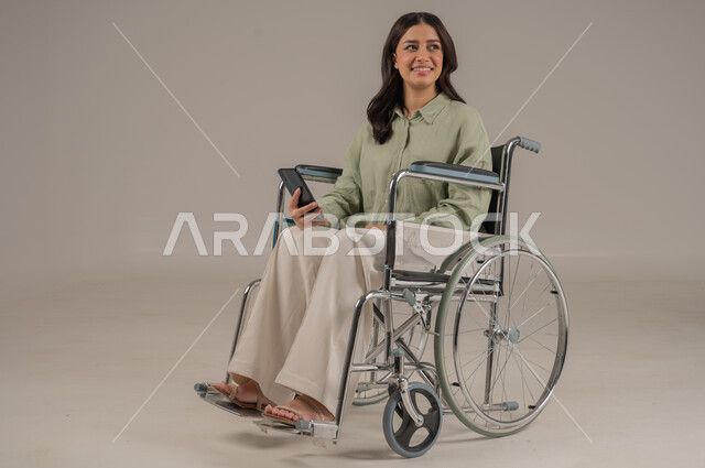 Chatting and communicating with family and friends, expressions of pleasure and happiness, using modern technical devices, portrait of a disabled Saudi Arabian Gulf woman wearing casual clothes sitting on a wheelchair holding a mobile phone in her hand, full body image, gray background