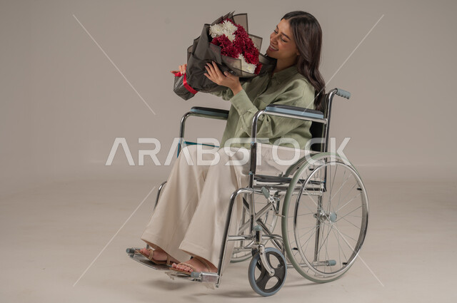 Natural flower shops and stores, gestures of joy and happiness, flower bouquet arrangement for holidays and happy occasions, portrait of a disabled Saudi Arabian Gulf woman wearing casual clothes sitting on a wheelchair holding a bouquet of roses in her hand, full body image, gray background