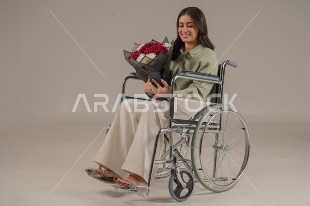 ايماءات السرور والسعادة، تنسيق باقات الزهور في الأعياد والمناسبات السعيدة، متاجر ومحلات بيع وشراء الأزهار الطبيعية، بورتريه لامراة عربية خليجية سعودية مقعد ترتدي الزي الكاجوال تجلس على كرسي متحرك تحمل بيدها باقة من الورود، صورة لكامل الجسم، خلفية رمادية