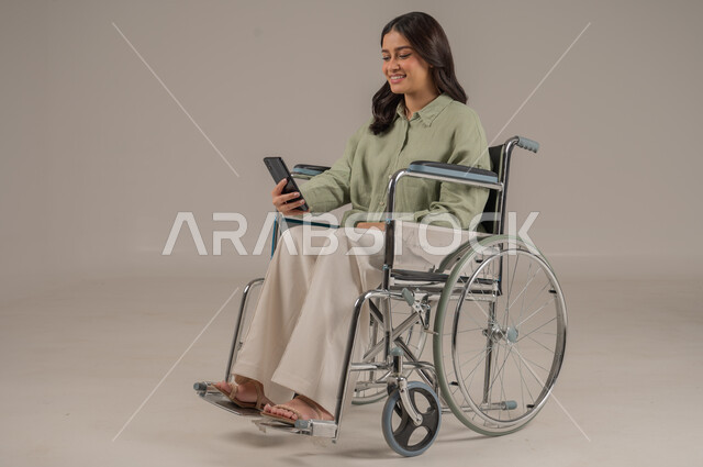 الدردشة والتواصل مع الاهل والاصدقاء، تعابير السرور والسعادة، استخدام الاجهزة التقنية الحديثة، بورتريه لامراة عربية خليجية سعودية مقعد ترتدي الزي الكاجوال تجلس على كرسي متحرك تحمل بيدها الهاتف المحمول، صورة لكامل الجسم، خلفية رمادية