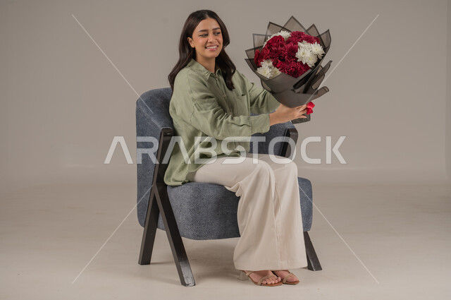 متاجر ومحلات بيع وشراء الأزهار الطبيعية، تعابير السرور والسعادة، تنسيق باقات الزهور في الأعياد والمناسبات السعيدة، بورتريه لامراة عربية خليجية سعودية ترتدي الزي الكاجوال تجلس على اريكة مريحة تحمل بيدها باقة من الورود، صورة لكامل الجسم، خلفية رمادية