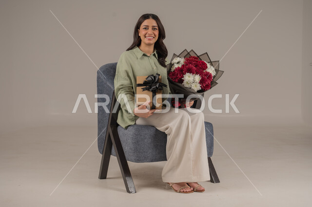متاجر ومحلات بيع وشراء الأزهار الطبيعية، تعابير السرور والسعادة، تنسيق باقات الزهور في الأعياد والمناسبات السعيدة، بورتريه لامراة عربية خليجية سعودية ترتدي الزي الكاجوال تجلس على اريكة مريحة تحمل بيدها باقة من الورود وصندوق هدايا، صورة لكامل الجسم، خلفية رمادية