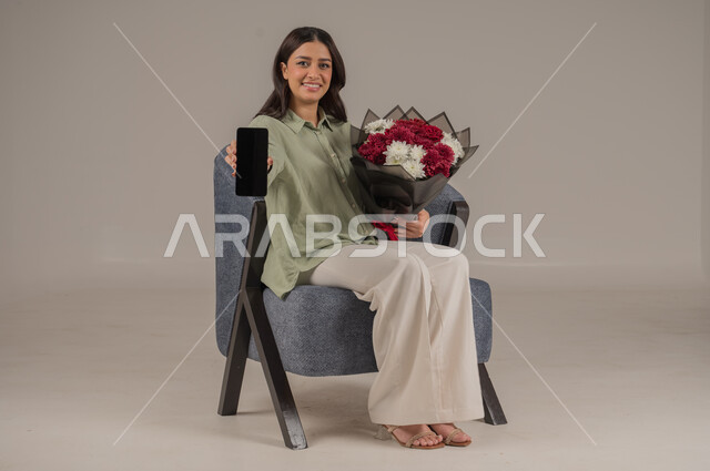 عرض شاشة سوداء فارغة، تنسيق باقات الزهور في الأعياد والمناسبات السعيدة، ايماءات السرور والسعادة، بورتريه لامراة عربية خليجية سعودية ترتدي الزي الكاجوال تجلس على اريكة مريحة تحمل بيدها باقة من الورود والهاتف المحمول، صورة لكامل الجسم، خلفية رمادية