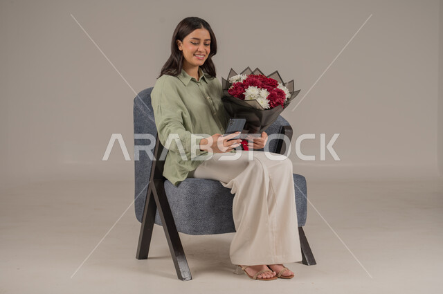 تنسيق باقات الزهور في الأعياد والمناسبات السعيدة، التقاط الصور التذكارية، ايماءات السرور والسعادة، بورتريه لامراة عربية خليجية سعودية ترتدي الزي الكاجوال تجلس على اريكة مريحة تحمل بيدها باقة من الورود والهاتف المحمول، صورة لكامل الجسم، خلفية رمادية