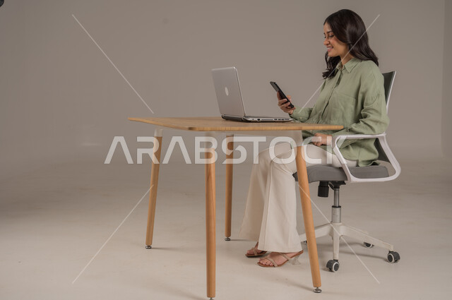 مهن ووظائف مكتبة، استخدام التقنيات الحديثة والمتطورة، انجاز مهام العمل عن بعد، بورتريه من الجانب لامراة عربية خليجية سعودية ترتدي الزي الكاجوال تجلس على طاولة المكتب تعمل على جهاز الهاتف المحمول، صورة لكامل الجسم، خلفية رمادية