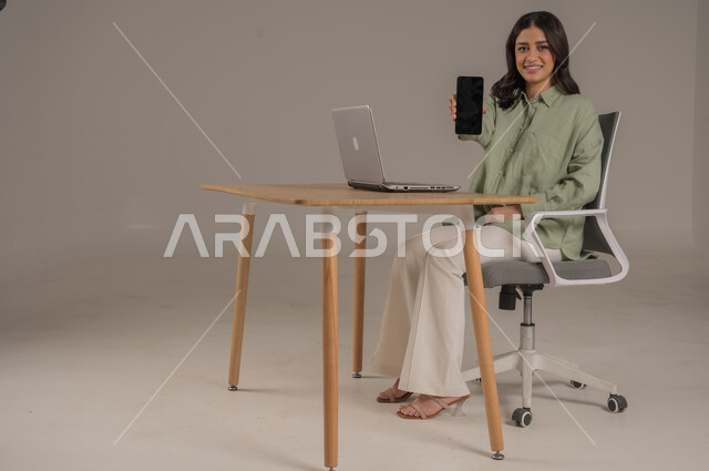عرض شاشة سوداء فارغة، انجاز العمل عن بعد، مهن ووظائف مكتبة، بورتريه لامراة عربية خليجية سعودية ترتدي الزي الكاجوال تجلس على طاولة المكتب تحمل بيدها الهاتف المحمول، صورة لكامل الجسم، خلفية رمادية