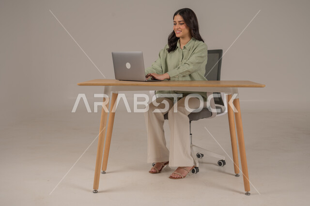 Using modern and advanced technologies, accomplishing work tasks remotely, office professions and jobs, portrait of a Saudi Arabian Gulf woman wearing casual clothes sitting at an office table working on a laptop, full body image, gray background