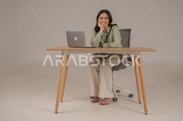  التفكير والتخطيط بالمستقبل، تعابيرالتمعن والتركيز، مفهوم التأمل والتفكر، إيجاد حل لمشكلة، بورتريه لامراة عربية خليجية سعودية ترتدي الزي الكاجوال تجلس على طاولة المكتب تضع يدها على وجهها بايماءات السرور، صورة لكامل الجسم، خلفية رمادية