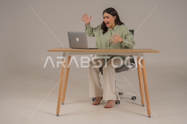 الشعور بالانزعاج والتوتر من سماع الأخبار السيئة والمزعجة، تعابير الدهشة والذهول، بورتريه لامراة عربية خليجية سعودية ترتدي الزي الكاجوال تجلس على طاولة المكتب ترفع يديها بإيماءات الصدمة والمفاجأة، صورة لكامل الجسم، خلفية رمادية