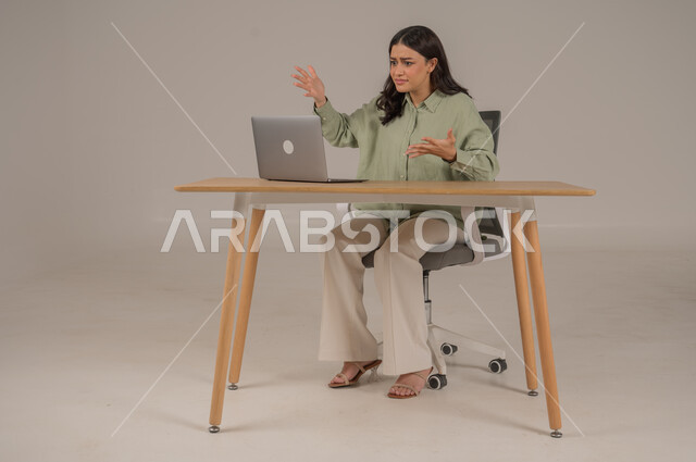 Expressions of surprise and astonishment, feeling upset and nervous from hearing bad and disturbing news, portrait of a Saudi Arabian Gulf woman wearing casual clothes sitting at the office table raising her hands with gestures of shock and surprise, full body image, gray background