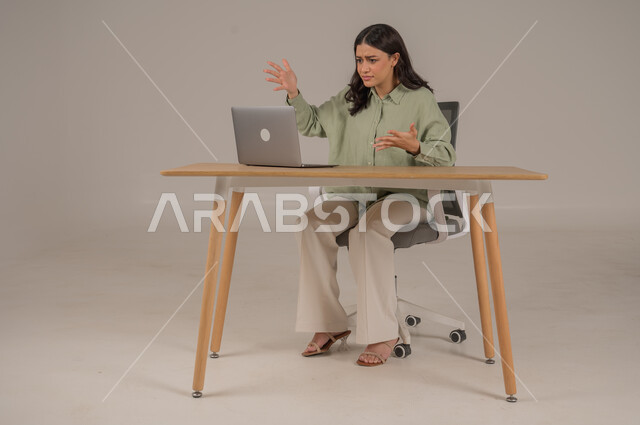 الشعور بالانزعاج والتوتر من سماع الأخبار السيئة والمزعجة، تعابير الدهشة والذهول، بورتريه لامراة عربية خليجية سعودية ترتدي الزي الكاجوال تجلس على طاولة المكتب ترفع يديها بإيماءات الصدمة والمفاجأة، صورة لكامل الجسم، خلفية رمادية