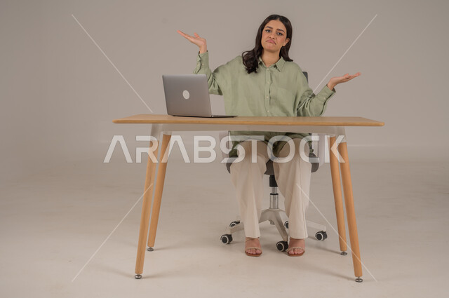 الشعور بالحيرة والجهل بأمر ما، التردد وانعدام المعرفة بخصوص شيء معين، استخدام التقنيات الحديثة والمتطورة، بورتريه لامراة عربية خليجية سعودية ترتدي الزي الكاجوال تجلس على طاولة المكتب ترفع يديها بإيماءات عدم التأكد، صورة لكامل الجسم، خلفية رمادية